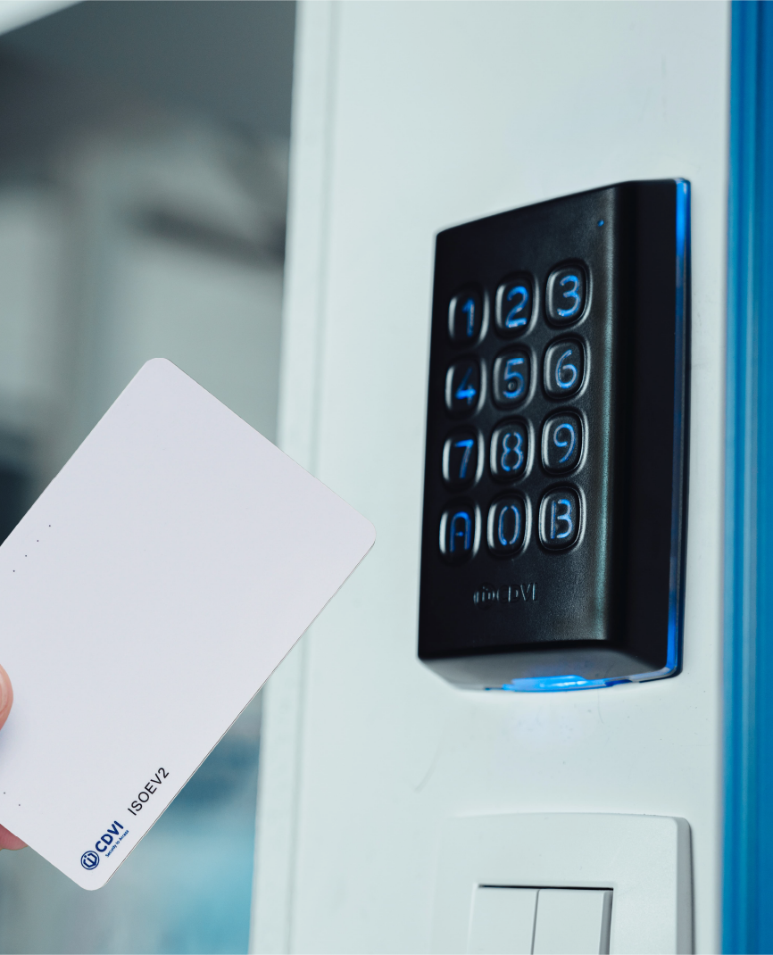 A black plastic keypad is mounted on a white wall with blue illuminated keys. A person's hand holding a white access card is reaching out to present the card to the keypad for access control validation